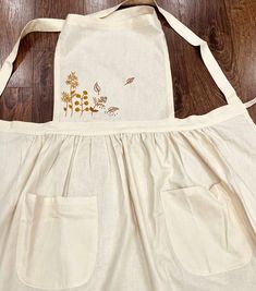 a white apron with embroidered flowers on the front and side pockets, sitting on a wooden floor