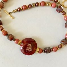 Beautiful handmade glass beads in a deep, rich burgundy red lying side by side with champagne and coral-colored beads remind me of a desert sunset.   Golden flecks sparkle in the center of the large glass disc bead.  Matching glass cubes flow up the neckband.  Gold-plated bead caps and spacers add elegance to the piece. Focal glass disc is 1 inch in diameter Glass and ceramic beads Gold-plated bead caps and spacers Gold-plated toggle clasp Necklace length:  15 3/4 inches Red Czech Glass Spiritual Jewelry, Red Czech Glass Jewelry With Spacer Beads, Brown Glass Beaded Necklaces As Gift, Brown Glass Beaded Necklaces For Gifts, Burgundy Beaded Necklaces With Round Beads As Gift, Red Murano Glass Jewelry With Round Beads, Handmade Burgundy Beaded Necklaces As Gift, Handmade Burgundy Beaded Necklaces For Gifts, Artisan Amber Beads For Gift