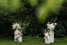 three white vases with flowers are sitting in the grass near some bushes and trees