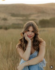 a woman sitting in the middle of a field with her hand on her chin and looking at the camera