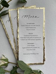 two menu cards with gold foil on them next to some greenery and flowers in front of a white wall