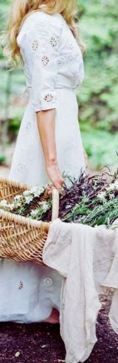 a woman in white dress holding a basket full of flowers next to a table covered with greenery