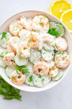 a bowl filled with shrimp and cucumbers next to lemon wedges on a white surface