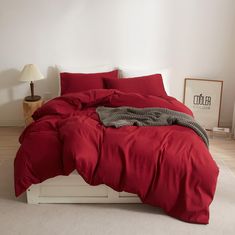 a bed with red sheets and pillows in a white room next to a lamp on the floor