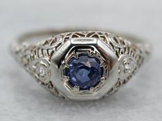 The height of Art Deco craftsmanship, this sapphire, and 18 karat white gold ring is set with bright diamonds, glittering and sparkling. Along with the detailed engraving work on all sides of the ring and the band, they are the perfect accent for the round cut and dark blue sapphire in the center. This gem has flashes of intense, saturated blue; a gorgeous combination with the white gold and diamonds. This can be worn as an engagement ring, an everyday ring, or just for special occasions!Metal: Vintage Blue Sapphire Ring With Diamond Accents, Blue Diamond Ring With Single Cut Diamonds, Blue Sapphire Ring With Single Cut Diamonds For Promise, Blue Sapphire Promise Ring With Single Cut Diamonds, Art Deco Blue Diamond Ring With Accents, Fine Jewelry Blue Sapphire Ring With Single Cut Diamonds, Formal Blue Rings With Single Cut Diamonds, Blue Art Deco Diamond Ring With Accents, Blue Diamond Ring With Single Cut Diamonds For Anniversary