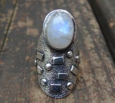 💙Handmade, Natural Rainbow Moonstone Sterling Silver Statement Ring Size 7 💙Featured in this listing is a sterling silver, handmade, natural stone ring. This lovely ring features a beautiful, Natural Rainbow Moonstone oval 14x11mm glowing stone. The stone has been bezel set into a handcrafted, unique setting made with all Sterling Silver. Detailed texturing, and rectangular and round accents, with oxidization, and a thick 8mm band. All details have been hand made. Gemstone: Natural Rainbow Moo Handmade Bohemian Moonstone Ring In Moon Shape, Handmade Bohemian Moonstone Moon Ring, Handmade Celestial Moonstone Ring In Moon Shape, Handmade Silver Moonstone Ring In Celestial Style, Handmade Moon-shaped Moonstone Ring In Sterling Silver, Handmade Unique Moonstone Ring, Unique White Moonstone Ring, Silver Mystical Moonstone Ring For Jewelry Making, Silver Moonstone Ring Stamped 925