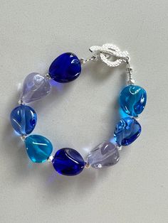 Another smooth glassy bracelet with beads by Samantha Capeling!  I made the bracelet with clear, smooth 15 x 13 mm nuggets, in a variety of ocean blues.  I accented them with 4 mm chunky Thai Hill Karen Tribe** sterling beads and a textured, but smooth, toggle clasp.  The bracelet measures about 7 3/4 inches long.  I have a 6 inch wrist and the bracelet fits me comfortably but not loosely.  In other words it doesn't drape down my hand. This is the exact bracelet you will be receiving, already ma Silver Beaded Bracelet, Sterling Silver Bead Bracelet, Ocean Colors, Hill Tribe Silver, Silver Bead Bracelet, Beaded Dangle Earrings, Sterling Silver Bead, Bracelet Gift, Matching Earrings