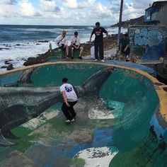 skateboarders are sitting on the edge of an empty pool by the ocean while others watch