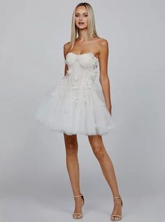 Embrace your inner romantic with this White Strapless Sweetheart Neck Corset Floral Appliqué Layered Tulle Mini Dress. This dreamy mini dress features a strapless sweetheart neckline, beautifully highlighting the décolletage for a feminine and elegant look. The corset bodice, with its tie-back detail, cinches the waist, offering a flattering silhouette and structured fit. Intricately designed floral appliqués adorn the bodice, adding a touch of whimsy and elegance. Layers of soft tulle create a Wedding Mini Dress With Sheer Bodice, Spring Bridesmaid Mini Corset Dress, Spring Mini-length Bridesmaid Corset Dress, Strapless Tulle Mini Dress For Spring, Fitted Tulle Mini Dress With Sweetheart Neckline, Chic Strapless Tulle Corset Dress, Spring Mini Dress With Sheer Bodice And Sweetheart Neckline, Sheer Mini Corset Dress For Summer, Sheer Mini Corset Dress For Spring