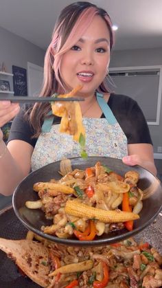 a woman holding a pan full of food with chopsticks in her hand and looking at the camera