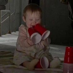 a small child sitting on the ground with a red cup in his mouth and one hand