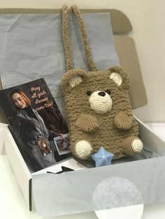 a crocheted teddy bear sitting in a box next to a book and a bag