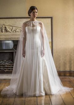 a woman in a wedding dress with a long veil on her head and shoulders, standing in front of a fireplace