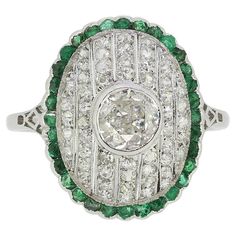 Here we have a fabulous emerald and diamond cluster ring crafted during pinnacle of the Art Deco movement. A single round faceted old European cut diamond sits slightly risen at the centre of the face in a full bezel setting. This principal stone proudly surmounts the concave backdrop consisting of 7 milgrain set channels filled with old cut diamonds which, in turn, are contained within an undulated border of perfectly matched emeralds around the outer edge. The piece is made complete by a ornate open under-gallery and split platinum band. Condition: Used (Very Good) Weight: 4.1 grams Ring Size: K (50) Face Dimensions: 18mm x 15mm Centre Diamond Weight: Approx. 0.50ct Diamond Details: Approx. Colour: G, Clarity: SI Total Remaining Diamond Weight: Approx. 0.55ct Total Emerald Dimensions: 32 Art Deco Oval Emerald Ring With Brilliant Cut, Heirloom Oval Emerald Ring With Single Cut Diamonds, Emerald Diamond Ring With Diamond Accents, Oval Emerald Ring With Single Cut Diamonds For Anniversary, Green Oval Diamond Ring With Single Cut, Green Oval Single Cut Diamond Rings, Classic Oval Emerald Ring With Single Cut Diamonds, Round Cluster Ring With Emerald And Diamond Accents, Vintage Green Diamond Platinum Ring