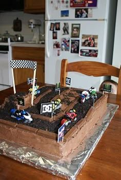 a birthday cake with dirt and race cars on it sitting on a table in front of a refrigerator