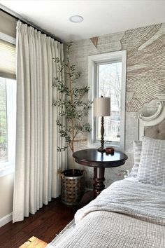 a bed sitting next to a window in a room with white curtains on the windowsill