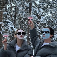 two women with face paint holding wine glasses in front of snow - covered pine trees