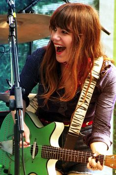 a woman with long hair playing guitar and singing into a microphone while standing in front of a green guitar