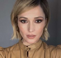 a close up of a person wearing a brown shirt and gold earrings with her hand on her shoulder