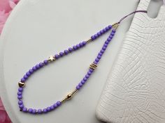 a purple beaded necklace next to a white cell phone case on a table with pink flowers