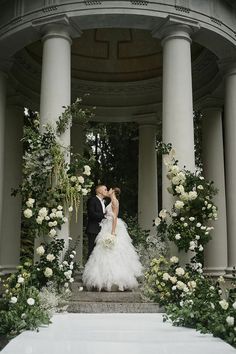 a bride and groom kissing in front of an archway with white flowers on the steps