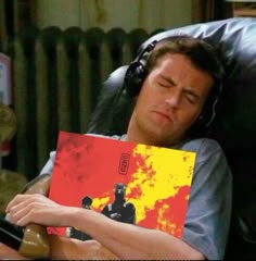 a man laying in a chair with headphones on while holding a red and yellow book