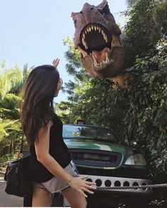 a woman standing in front of a fake dinosaur head next to a car with it's mouth open