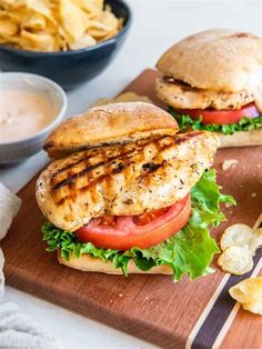 two chicken burgers with lettuce and tomatoes on a cutting board next to chips