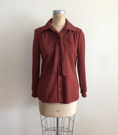 "Long sleeved, dark red/maroon, heavyweight blouse with neck tie, from the 1970s. Bust measures 20.5\" (52 cm) flat across and waist measures 18.5\" (47 cm) flat across. The blouse is 28\" (71 cm) in length. Polyester. Original size is a 14. Tie is removable." Red Collared Blouse For Fall, Classic Fitted Brown Blouse, Classic Red Blouse For Fall, Vintage Fitted Brown Blouse, Vintage Red Blouse For Fall, Vintage Burgundy Tops For Fall, Retro Fall Office Tops, Red Collared Vintage Blouse, Vintage Red Collared Blouse