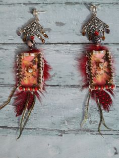 two earrings with red feathers and beads hanging from hooks on a wooden surface, one is decorated with an image of a woman's face