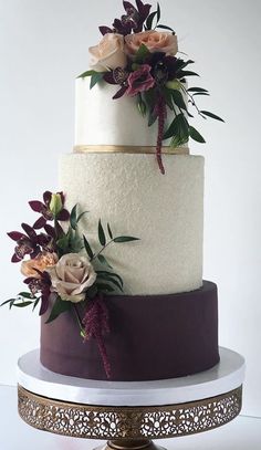 a three tiered wedding cake with purple and white flowers on the top, sitting on a gold stand