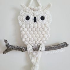 a crocheted white owl hanging on a tree branch with eyes wide open and one eye partially closed