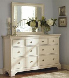 a white dresser with flowers on top and a mirror in the corner above it, against a blue wall