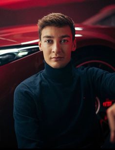 a young man sitting in front of a red car with his hand on the steering wheel