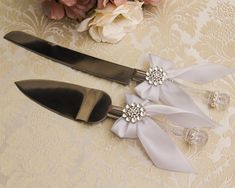 two wedding cake knifes with bows and crystal beads on them sitting next to flowers