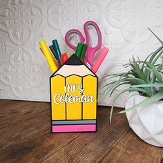 a pencil holder with scissors and markers in it on a wooden table next to a potted plant