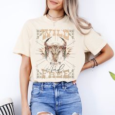 Embrace your untamed spirit with our captivating T-shirt design, a fusion of Western ruggedness and Boho charm. A majestic cow skull, intricately adorned with vibrant Boho motifs, rests upon a bed of ethereal white blooms and lush foliage, illuminated by the warmth of a Boho sun. Above, the word 'Wild' arcs boldly in retro font, while 'And Free' gracefully follows below in script, encapsulating the essence of freedom and wanderlust. Elevate your style with this symbol of untamed beauty, perfect Fall Festival Graphic Print T-shirt, Bohemian Short Sleeve T-shirt For Fall, Free-spirited Short Sleeve Cotton T-shirt, Summer Free-spirited Short Sleeve T-shirt, Free-spirited Short Sleeve Spring T-shirt, Hippie Graphic Print T-shirt For Fall, Free-spirited Graphic Print T-shirt For Summer, Free-spirited Cotton T-shirt With Graphic Print, Free-spirited Cotton Crew Neck T-shirt