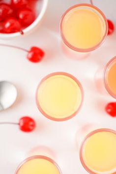 four glasses filled with yellow liquid and cherries next to a bowl of cherries