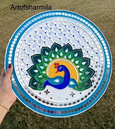 a hand holding a plate with a peacock painted on it in the middle of grass