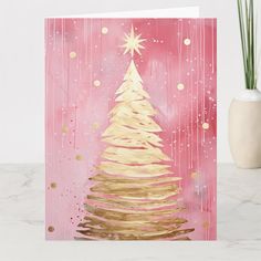 a pink and gold christmas card with a white tree on the front, against a pink background