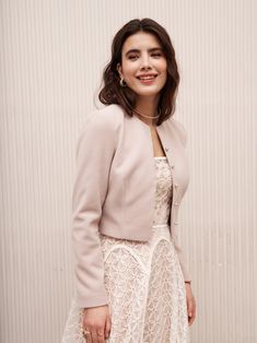 a woman standing in front of a wall wearing a white dress and jacket with lace detailing