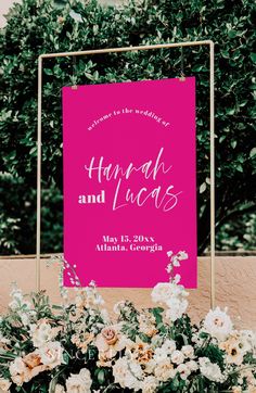 a pink and gold wedding sign with white flowers in the foreground is surrounded by greenery