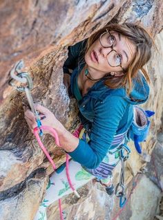 a woman climbing up the side of a rock with her hands in her pockets and wearing glasses