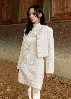 a woman in a white dress and jacket standing on dirt ground next to a building