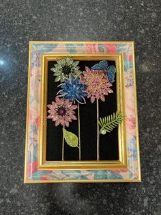 some flowers are in a gold frame on a black counter top and there is a butterfly flying over them