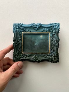 a person holding up a miniature painting in front of a white wall with a blue frame