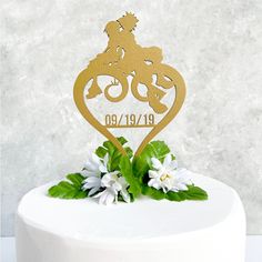 a white cake topped with a gold wedding cake topper next to flowers and greenery