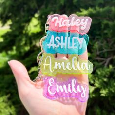 a hand holding three personalized keychains in front of some trees and bushes