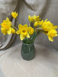 a green vase filled with yellow flowers on top of a white cloth covered tablecloth