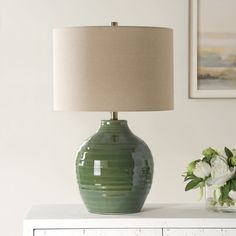 a green vase sitting on top of a white table next to a lamp and flowers